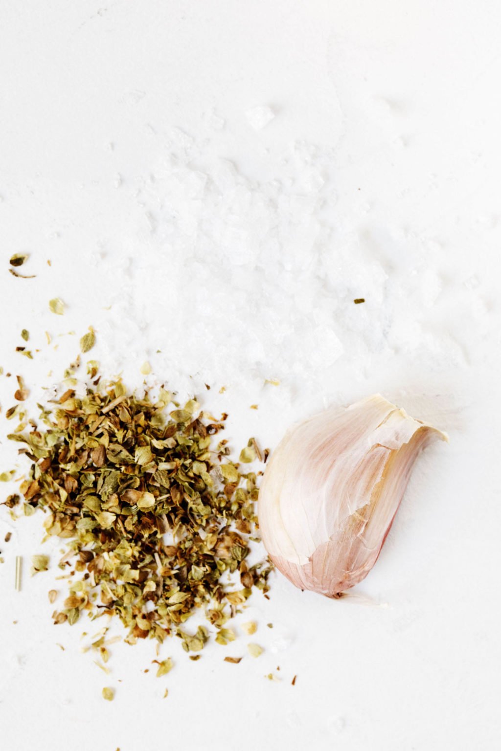 Dried oregano and an unpeeled clove of garlic rest on a white surface.