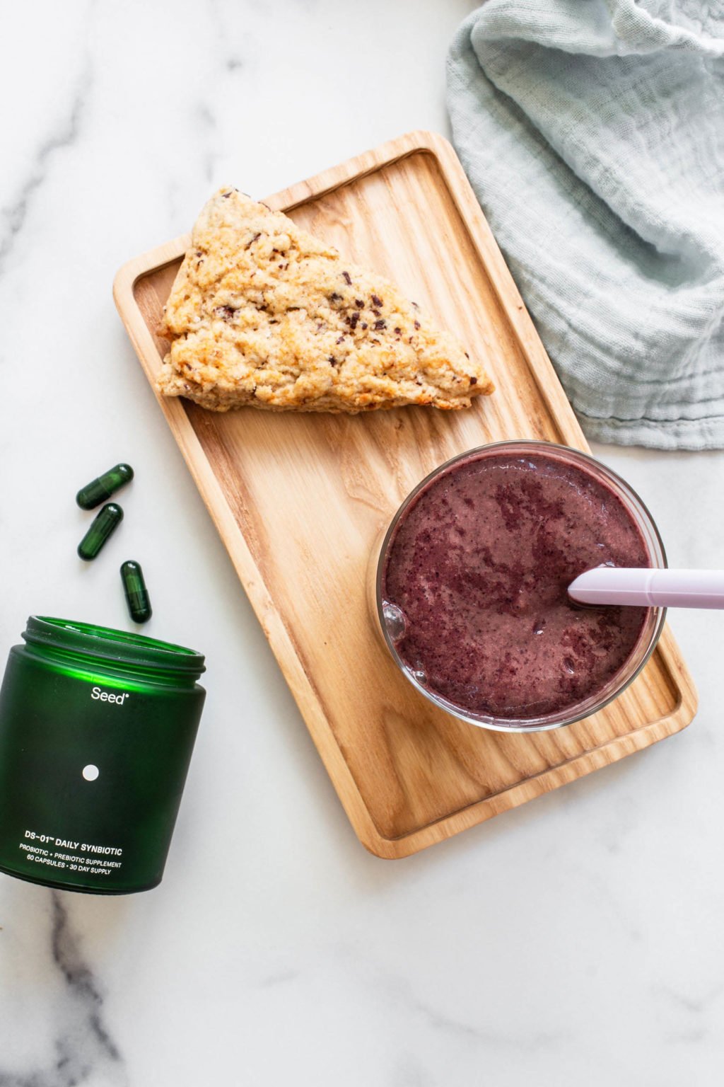 An overhead image of a purple smoothie, which is arranged on a small wooden board with a single scone. A bottle of pills and a cloth napkin are nearby.