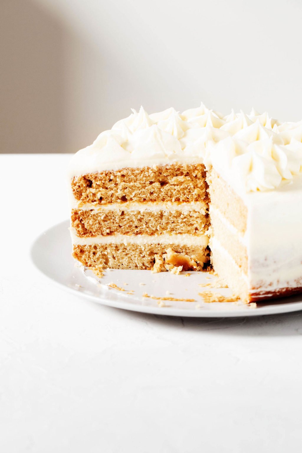 A three layered vanilla cake rests on a round plate. The cake has had a slice cut into it and is decorated with white buttercream frosting.