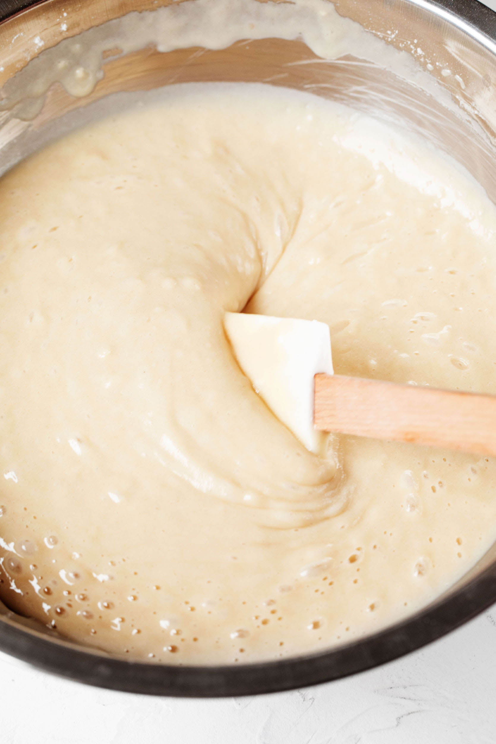 A mixing bowl has been filled with vanilla cake batter, which is being stirred with a spatula.