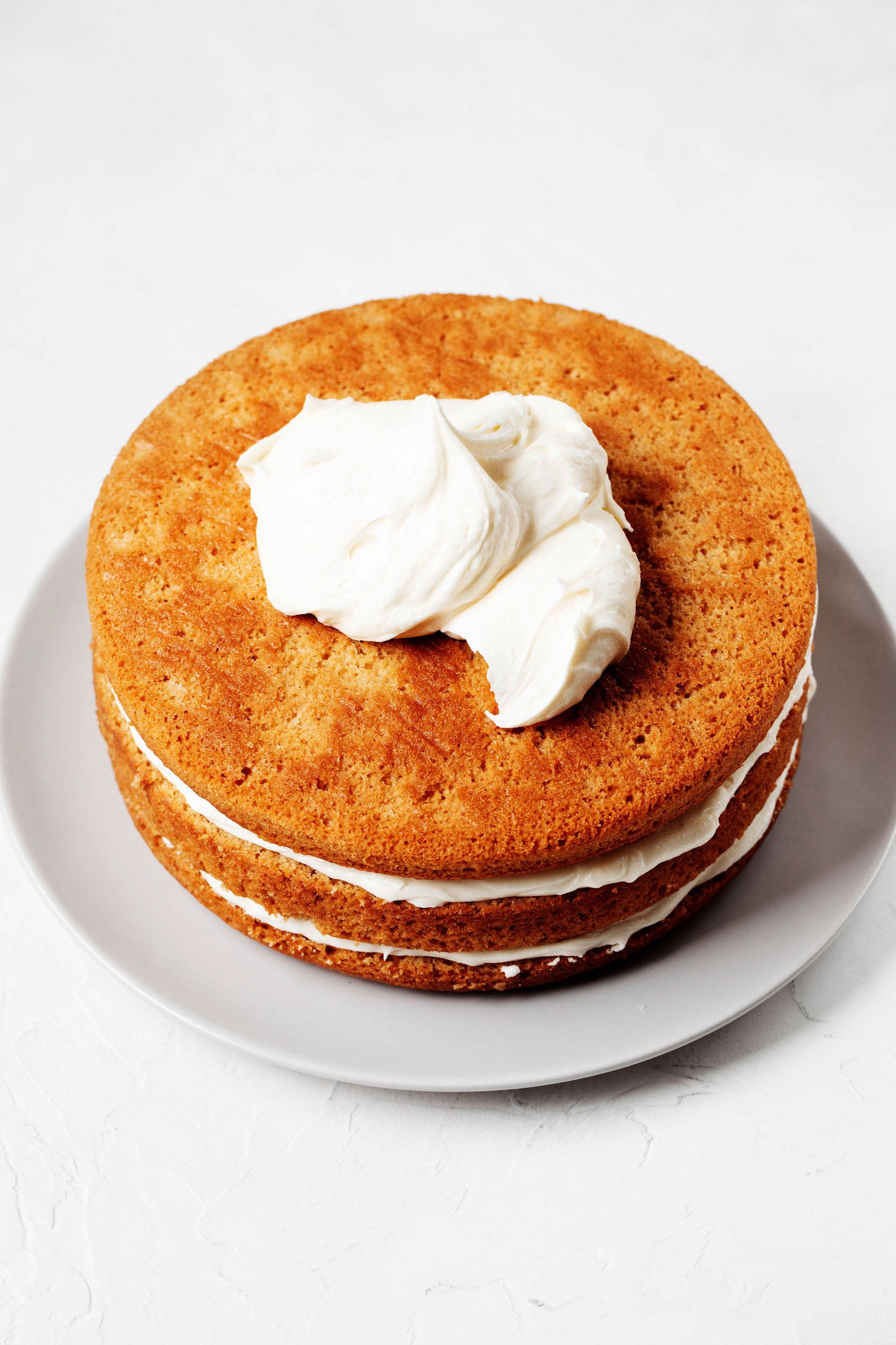 Three layers of vanilla cake have been layered with white, vanilla frosting. The cake is being decorated on a white, round plate.