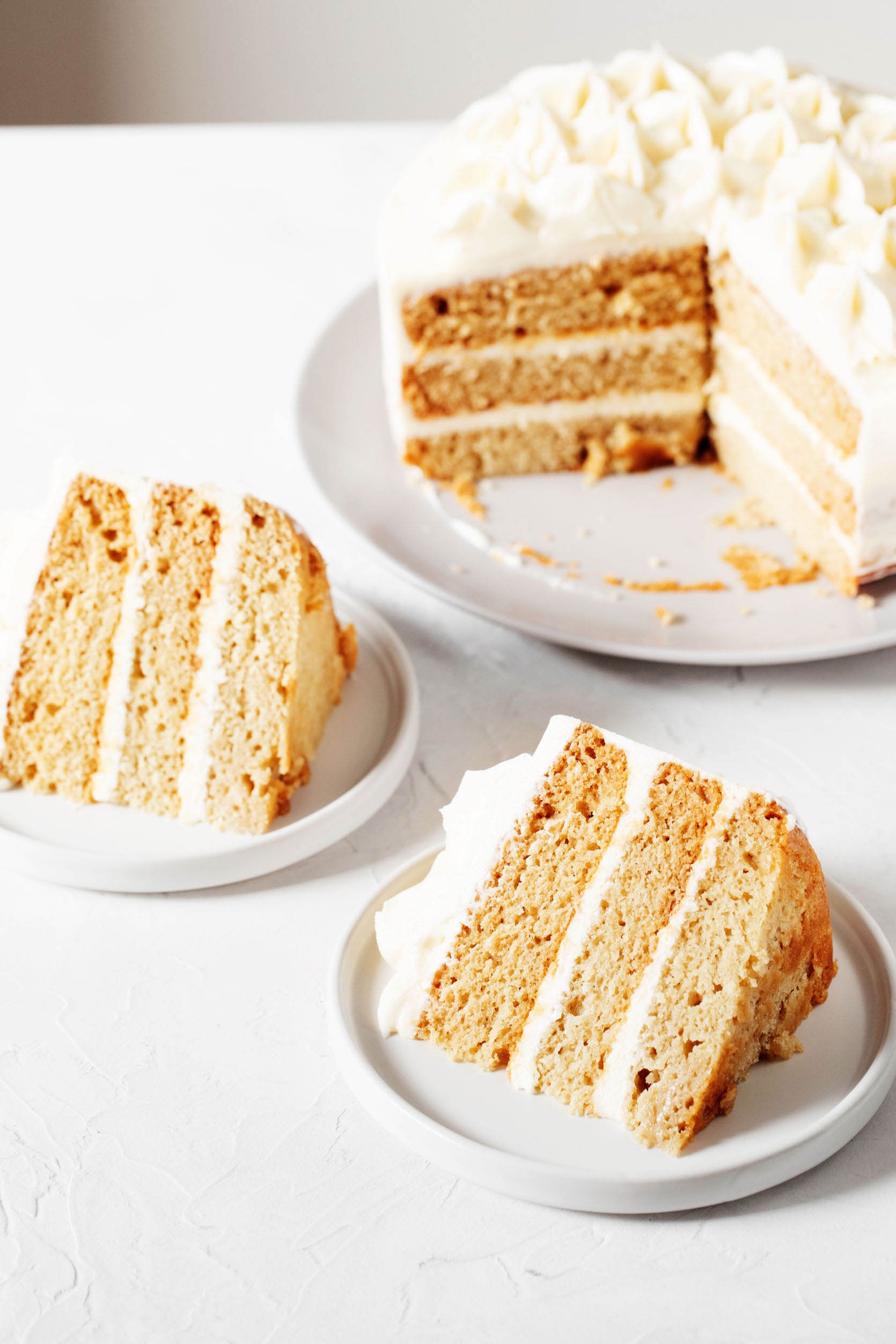 A pale yellow layer cake with whipped, white buttercream frosting has had slices cut out. Two slices rest on small dessert plates on top of the cake, all on top of a white surface.
