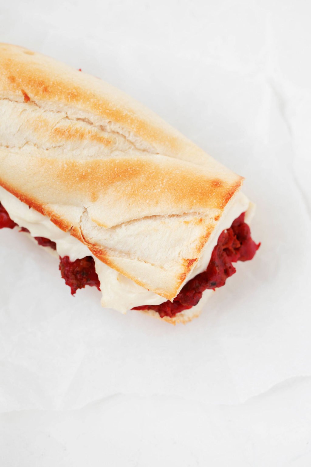 An overhead image of sliced Italian bread, which has been piled with red sauce and a creamy cheese sauce. It rests on a piece of parchment paper.