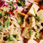 A zoomed in, overhead photograph of a tangle of pad thai noodles, tofu, vegetables, and herbs.