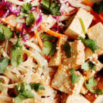 A zoomed in, overhead photograph of a tangle of pad thai noodles, tofu, vegetables, and herbs.