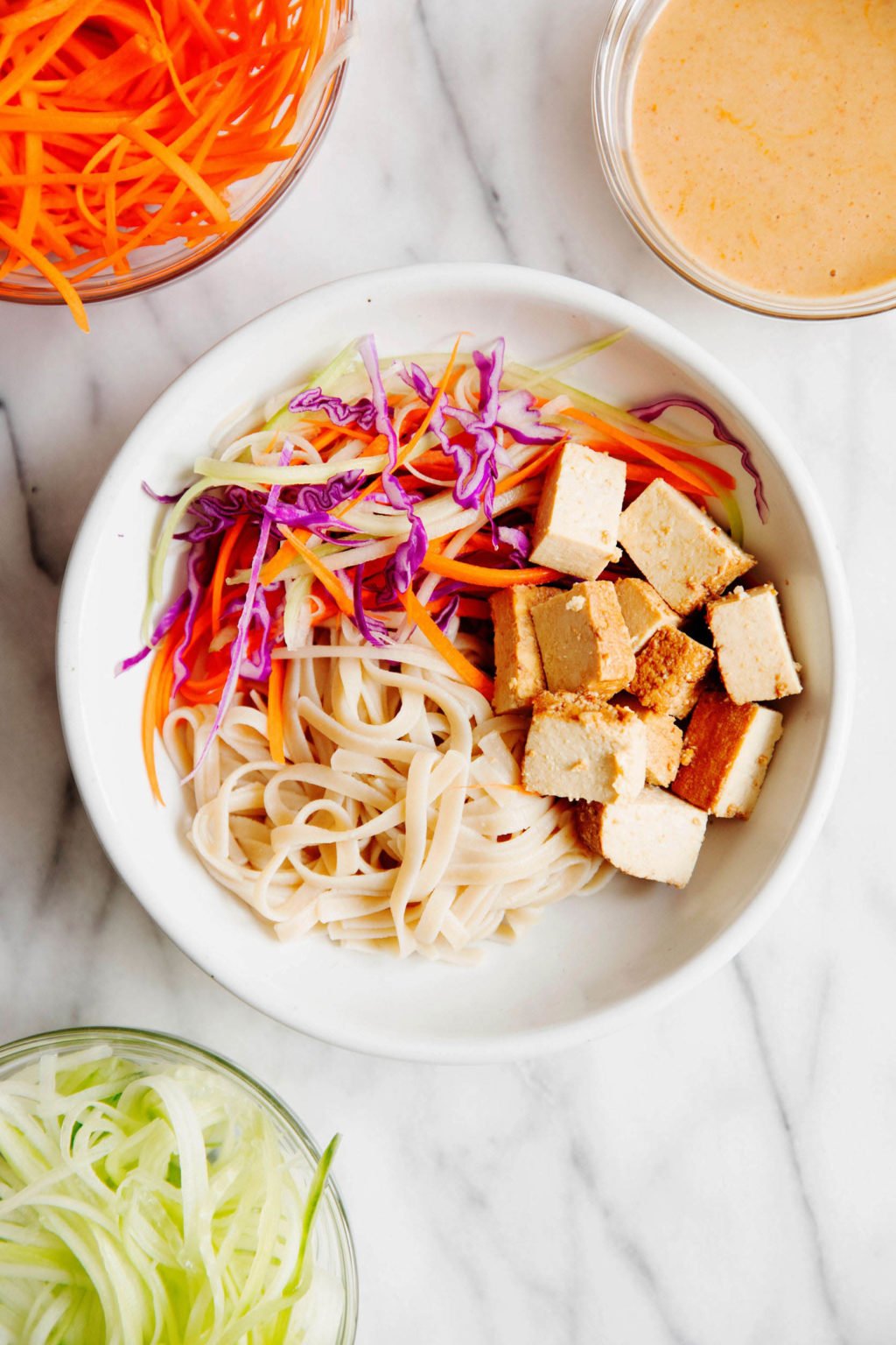 A bowl of tofu, noodles, and vegetables, surrounded by small glass bowls with prepared ingredients in them. The bowls rest on white marble.