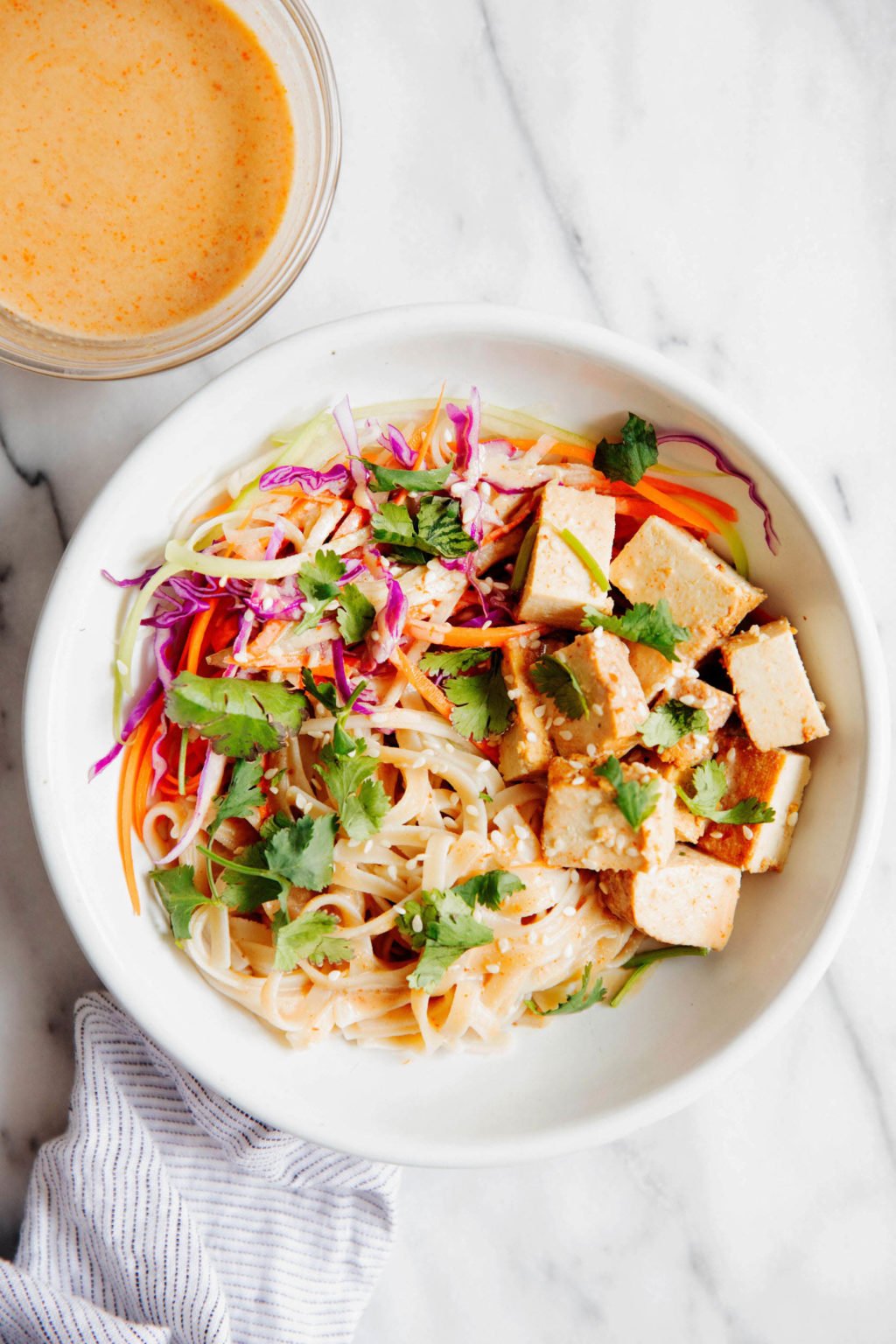 A bright teriyaki tofu noodle bowl is resting on white marble, with a small bowl of sauce nearby.