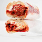 A vegan tempeh meatball sub is wrapped in parchment paper and resting on a white surface.