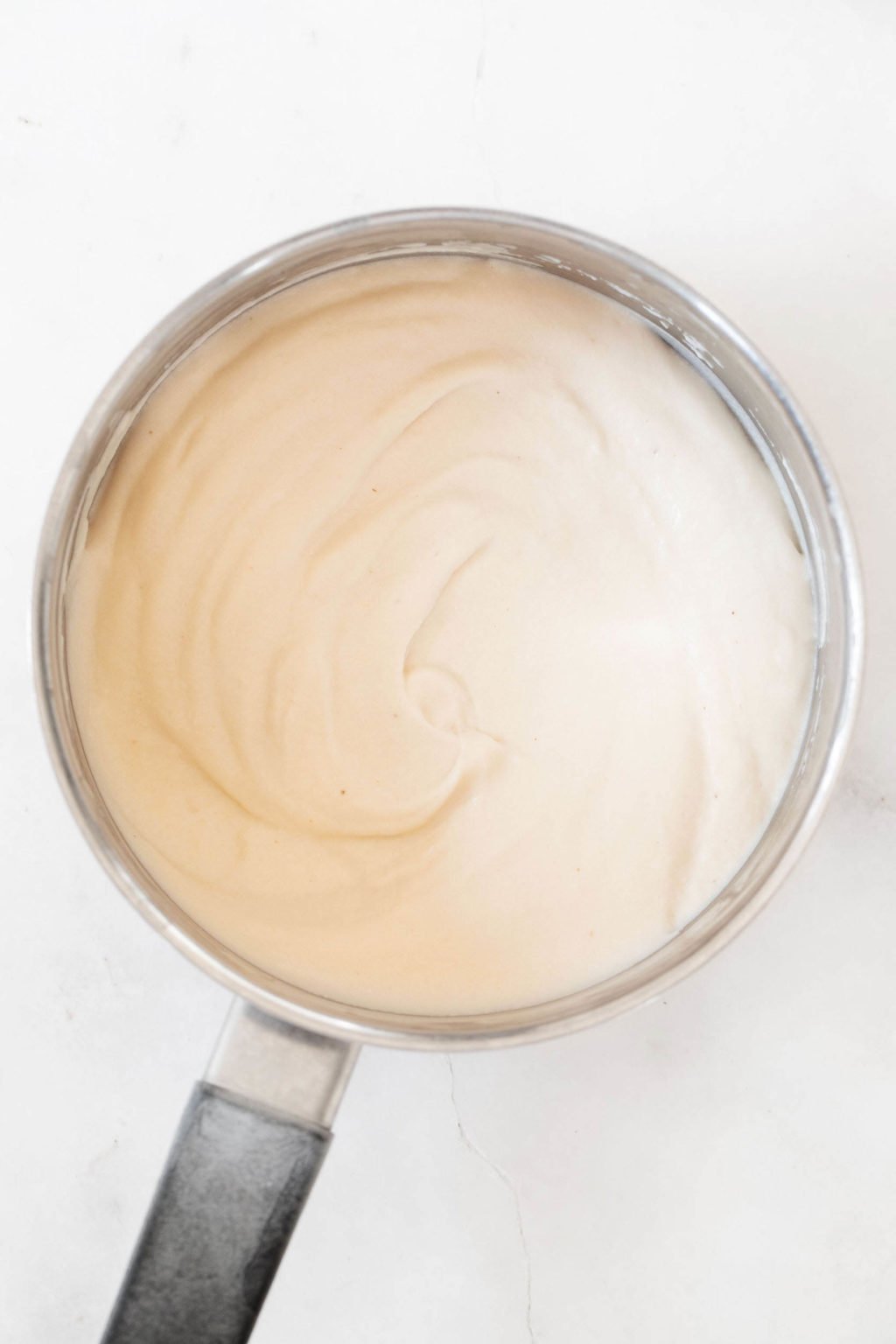 An overhead image of a silver saucepan with a black handle. The saucepan is full of a creamy, rich white sauce.