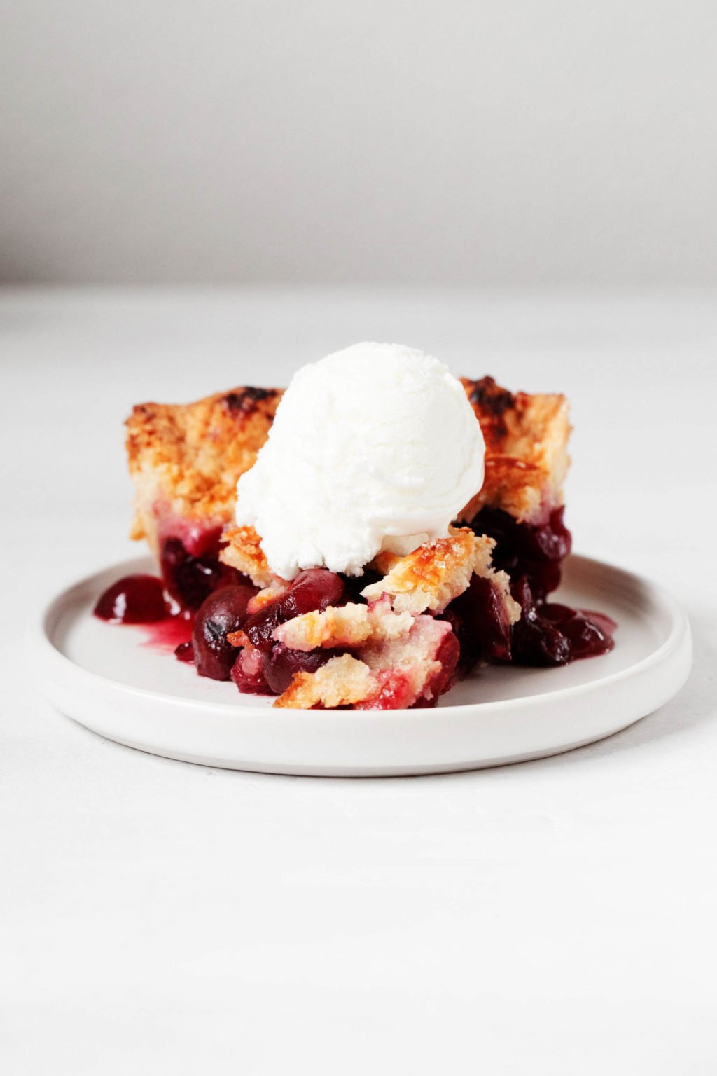 A slice of rustic, fruit dessert rests on a small, round white dessert plate. The flaky dessert is topped with a white scoop of vanilla ice cream.