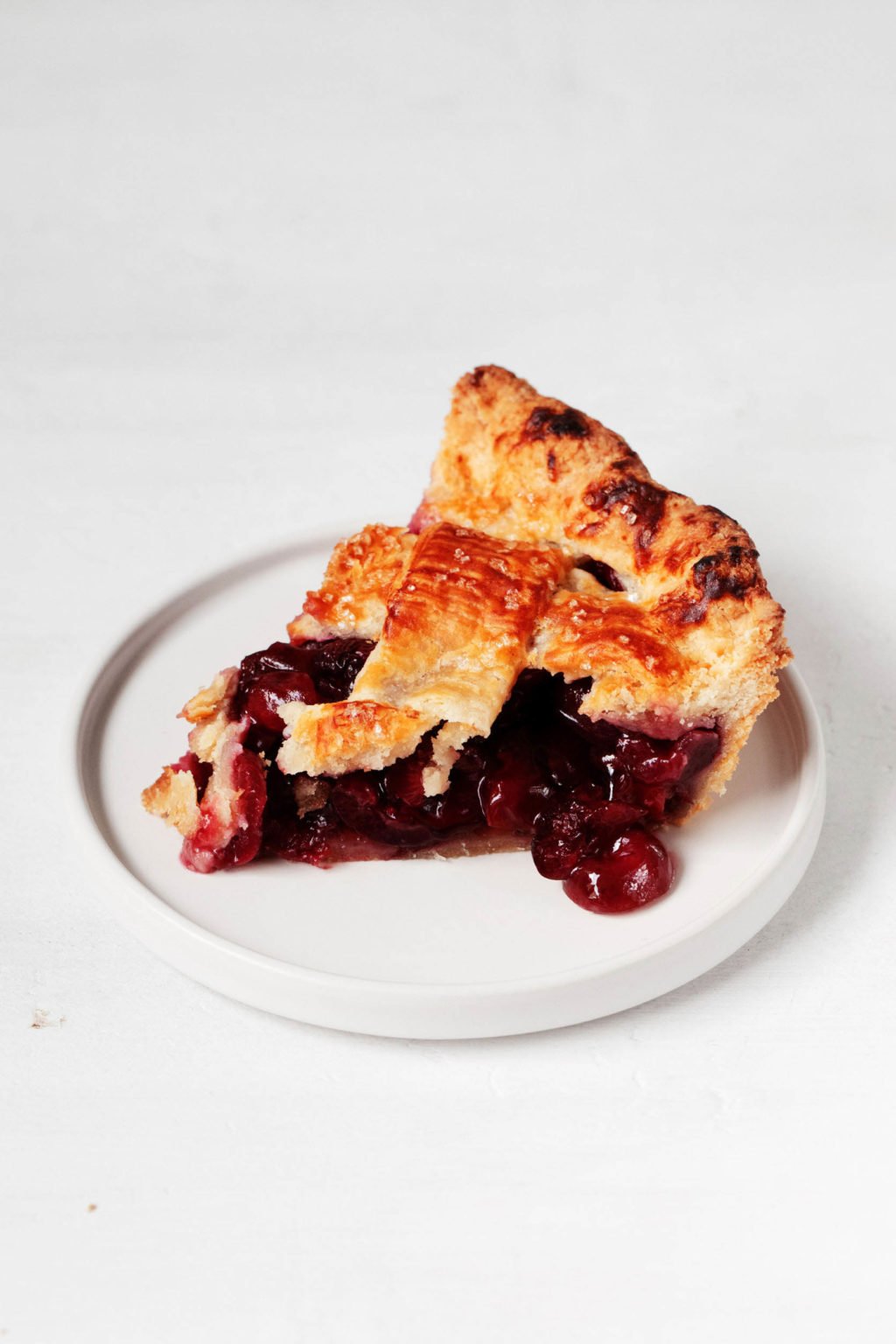 A slice of cherry pie rests on a small, white dessert plate. It's filled with juicy cherries and has a lattice top crust.