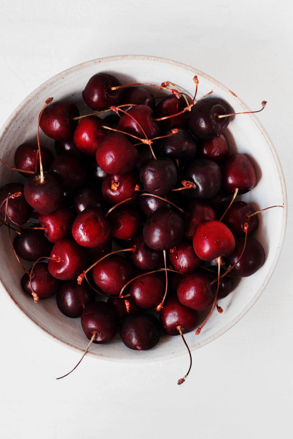 A bowl of whole, dark red sweet cherries. The white bowl is pictured against a white backdrop.