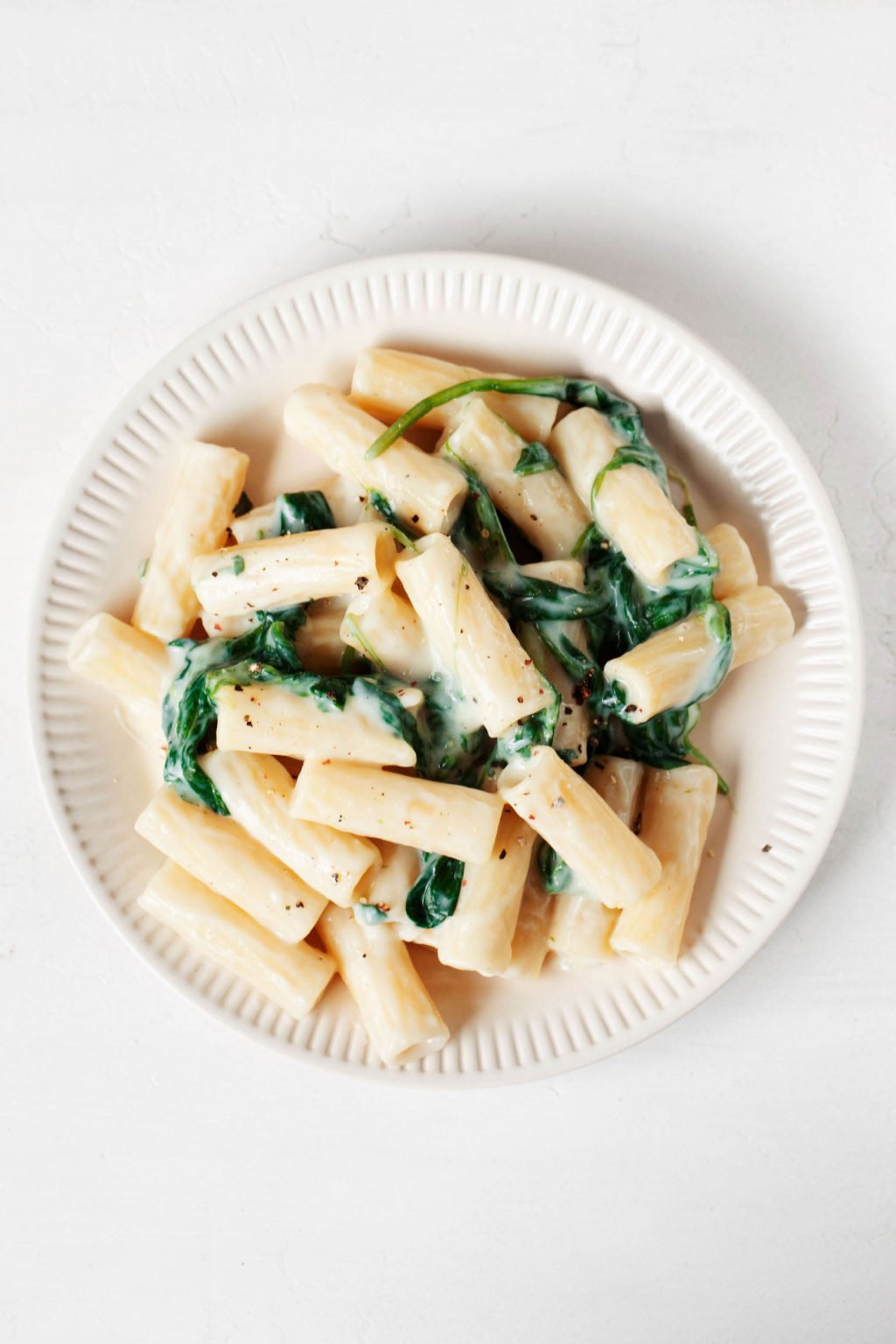 An overhead image of a rimmed, cream colored ceramic plate on a cream colored surface. The plate holds a creamy plant-based pasta dish with greens.