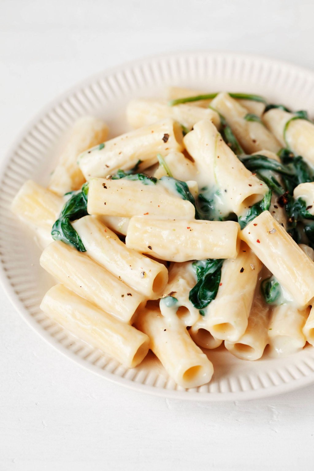 A rimmed, cream colored ceramic dish holds a creamy pasta dish with spinach.