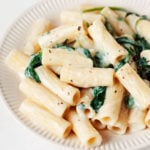 A rimmed, cream colored ceramic dish holds a creamy pasta dish with spinach.