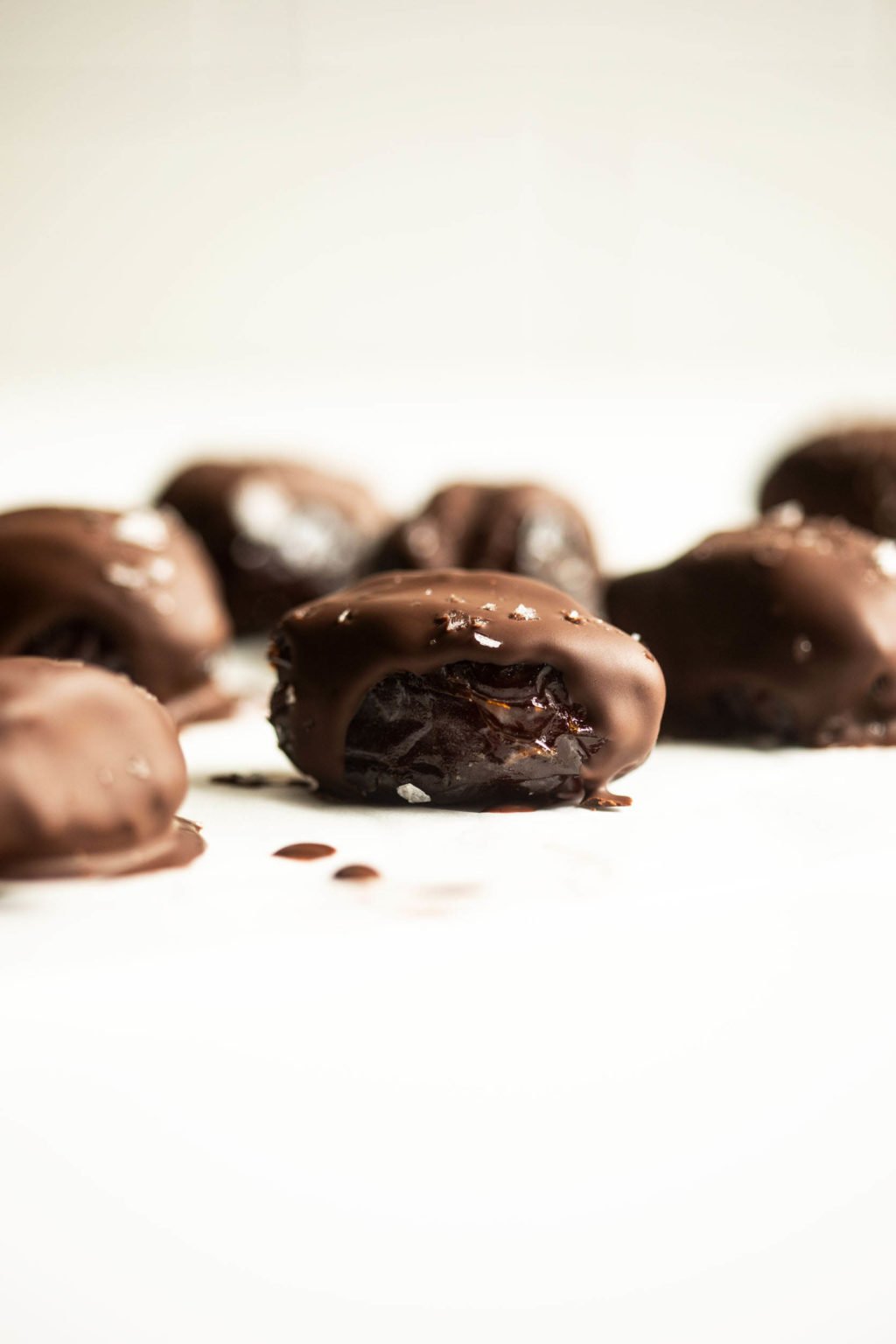 Medjool dates that have been drizzled with dark chocolate and sea salt are arranged on a white surface.