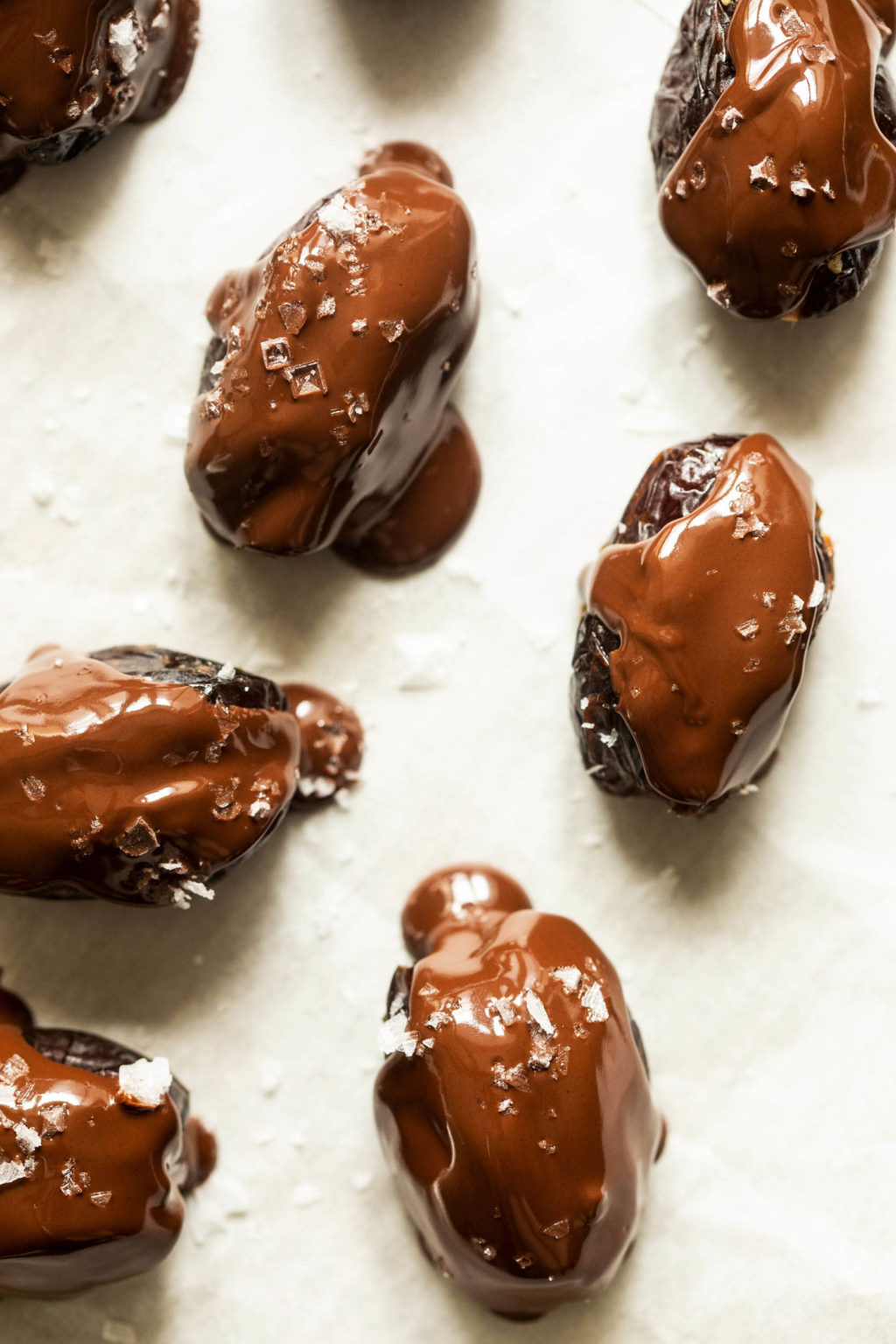 Chocolate dipped, stuffed dates have been sprinkled with flaky sea salt on a sheet of white parchment paper.