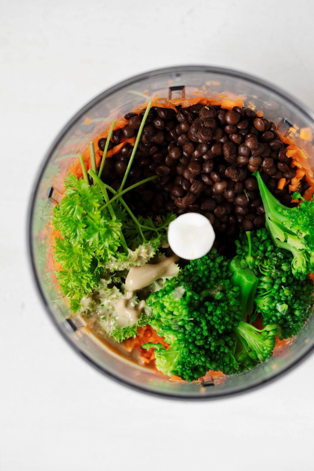 The bowl of a food processor is filled with lentils, broccoli, carrots, and herbs.