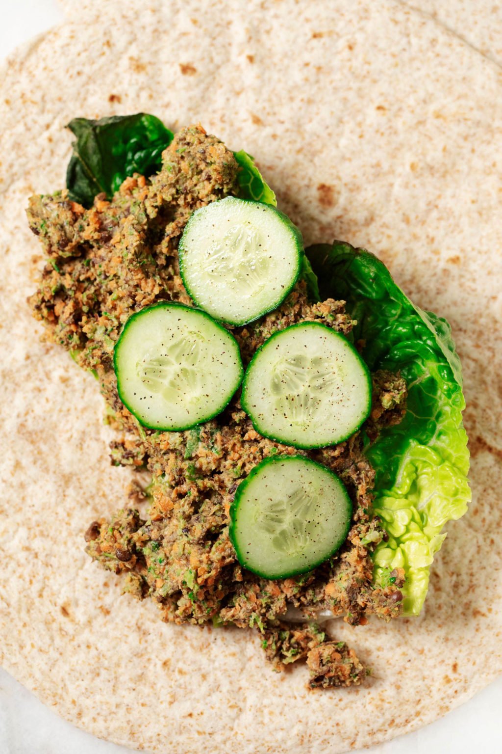 A whole grain wrap is being filled with a mixture of lentils and vegetables and some cucumber slices.
