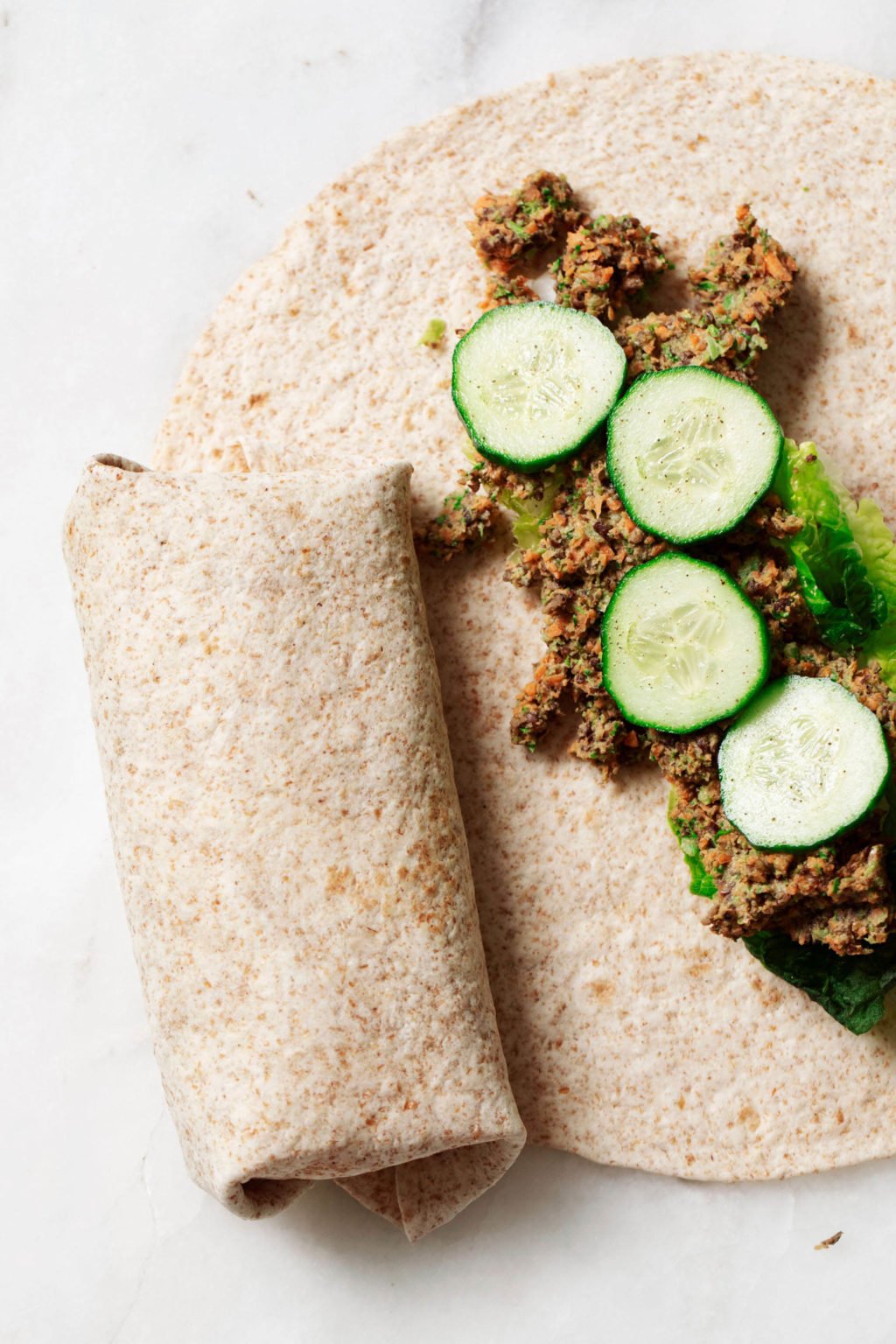 Lentil tahini wraps are being prepared on a white work surface. They're stuffed with cucumber slices and a lentil vegetable filling.