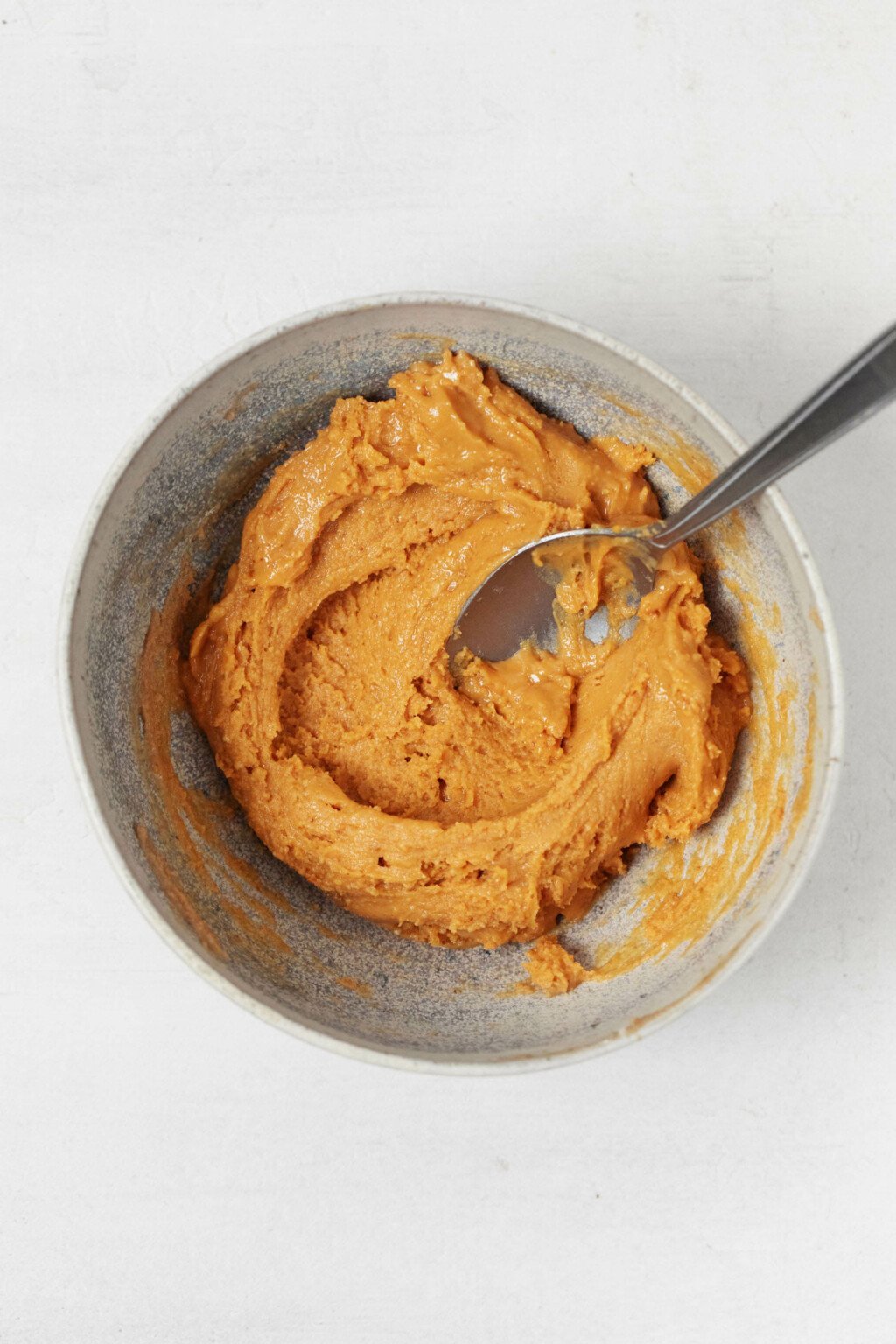 A small, ceramic bowl holds a mixture of peanut butter and protein powder, which is being stirred with a spoon.