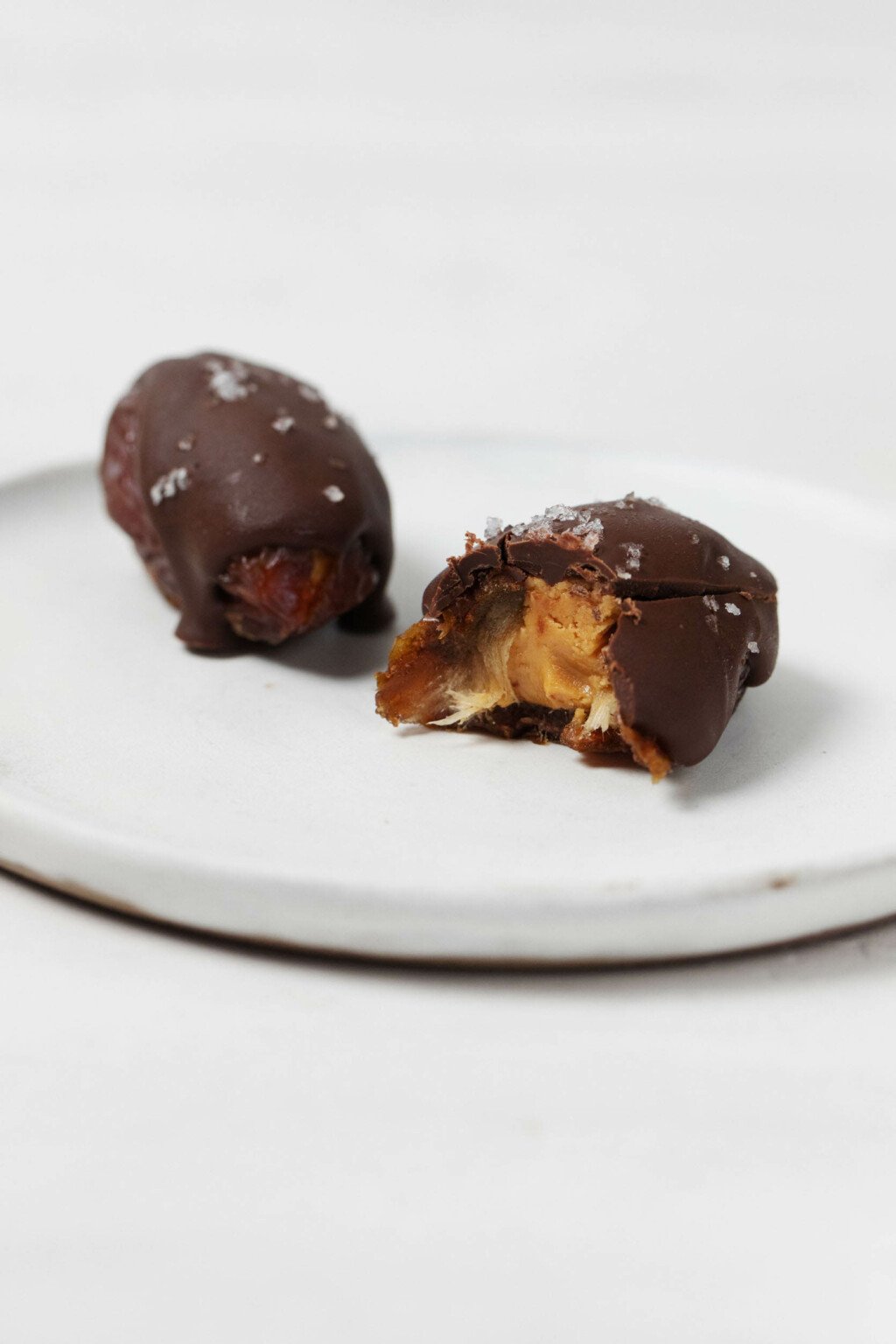 A peanut butter stuffed Medjool date has been topped with chocolate and sea salt. It rests on a white plate.