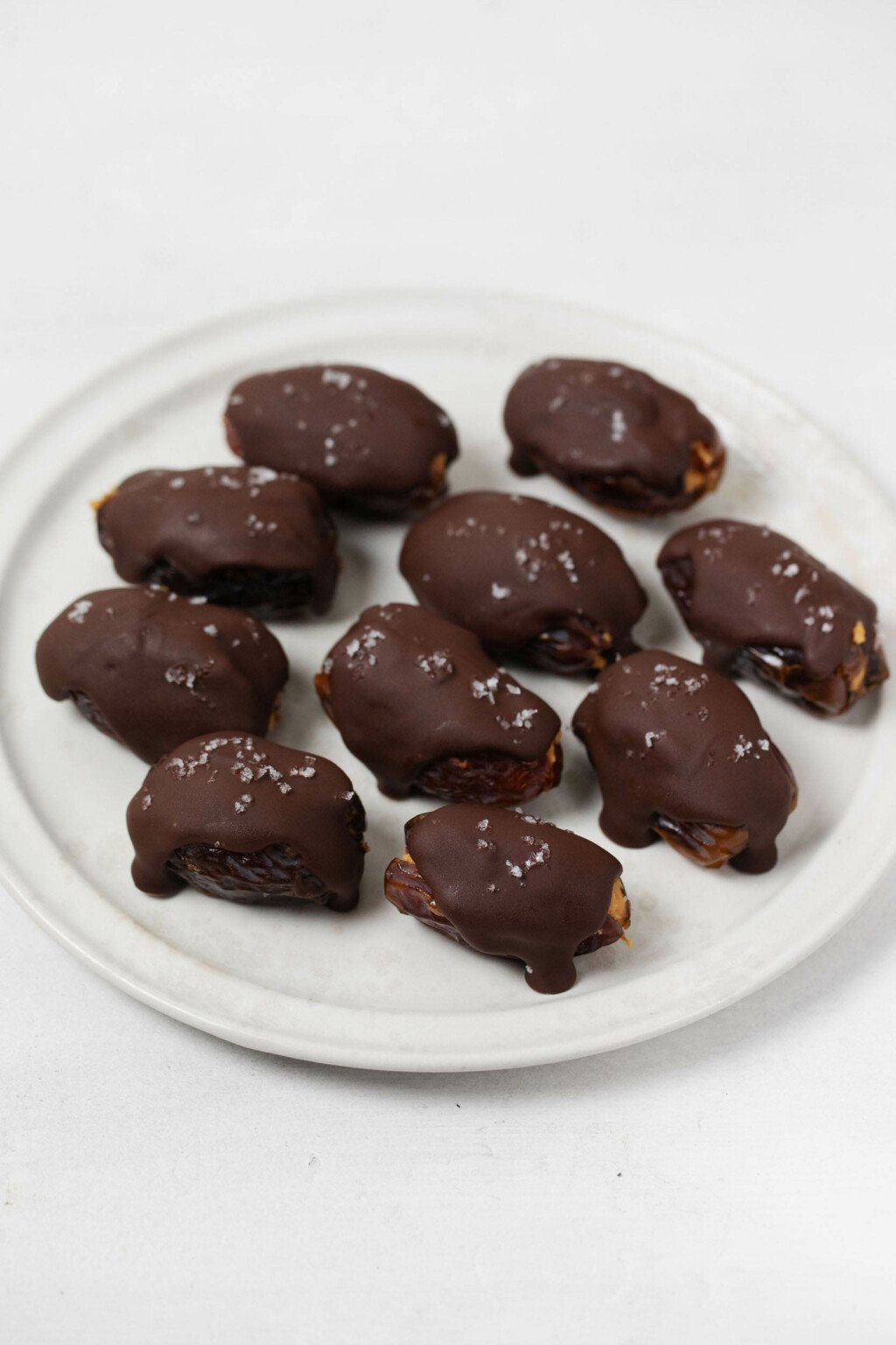 Medjool dates are lined up on a round white plate. They are all dipped in dark chocolate and sprinkled with sea salt.