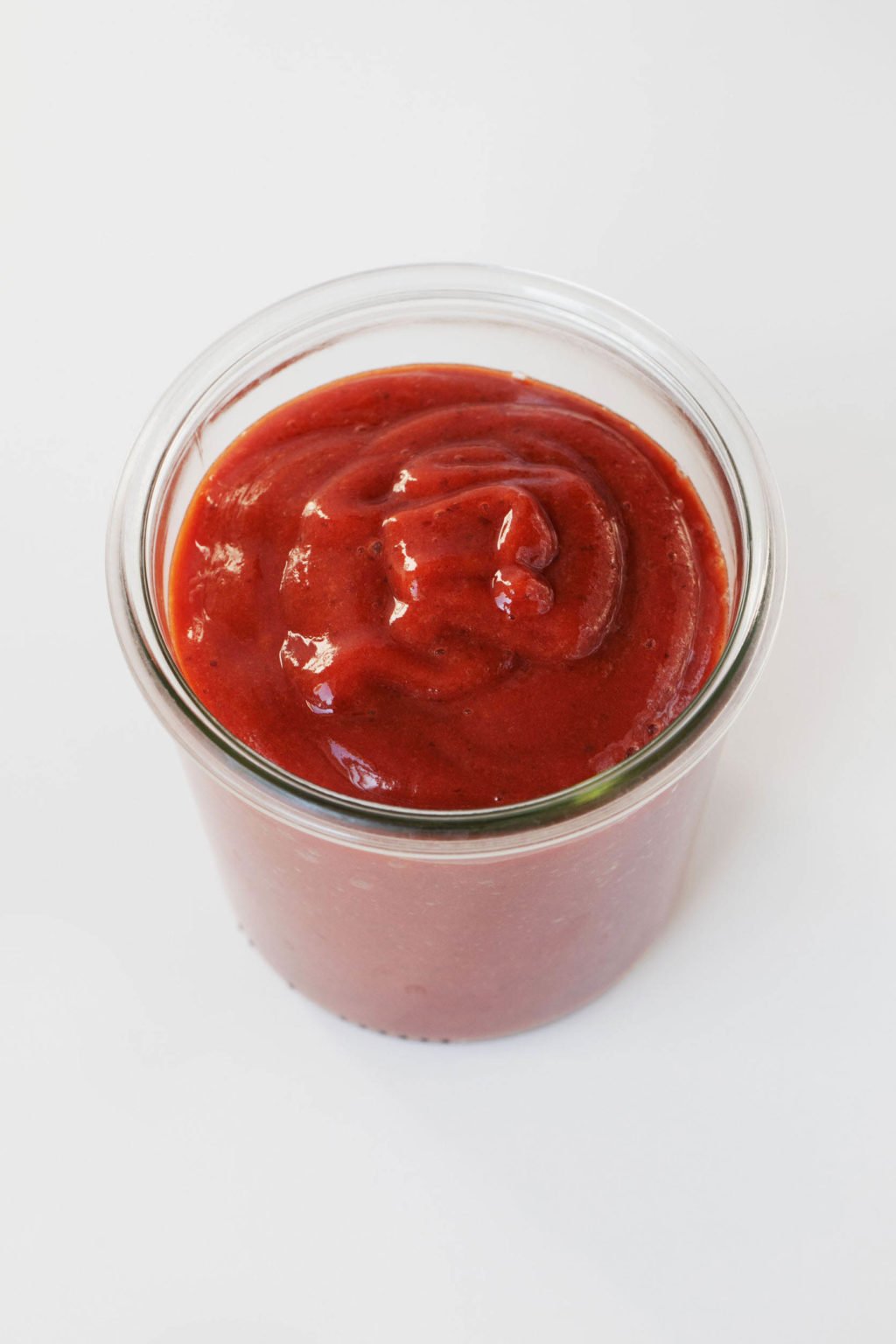 An overhead image of a red orange, vegan date BBQ sauce. It is held in a glass jar and rests on a while surface.