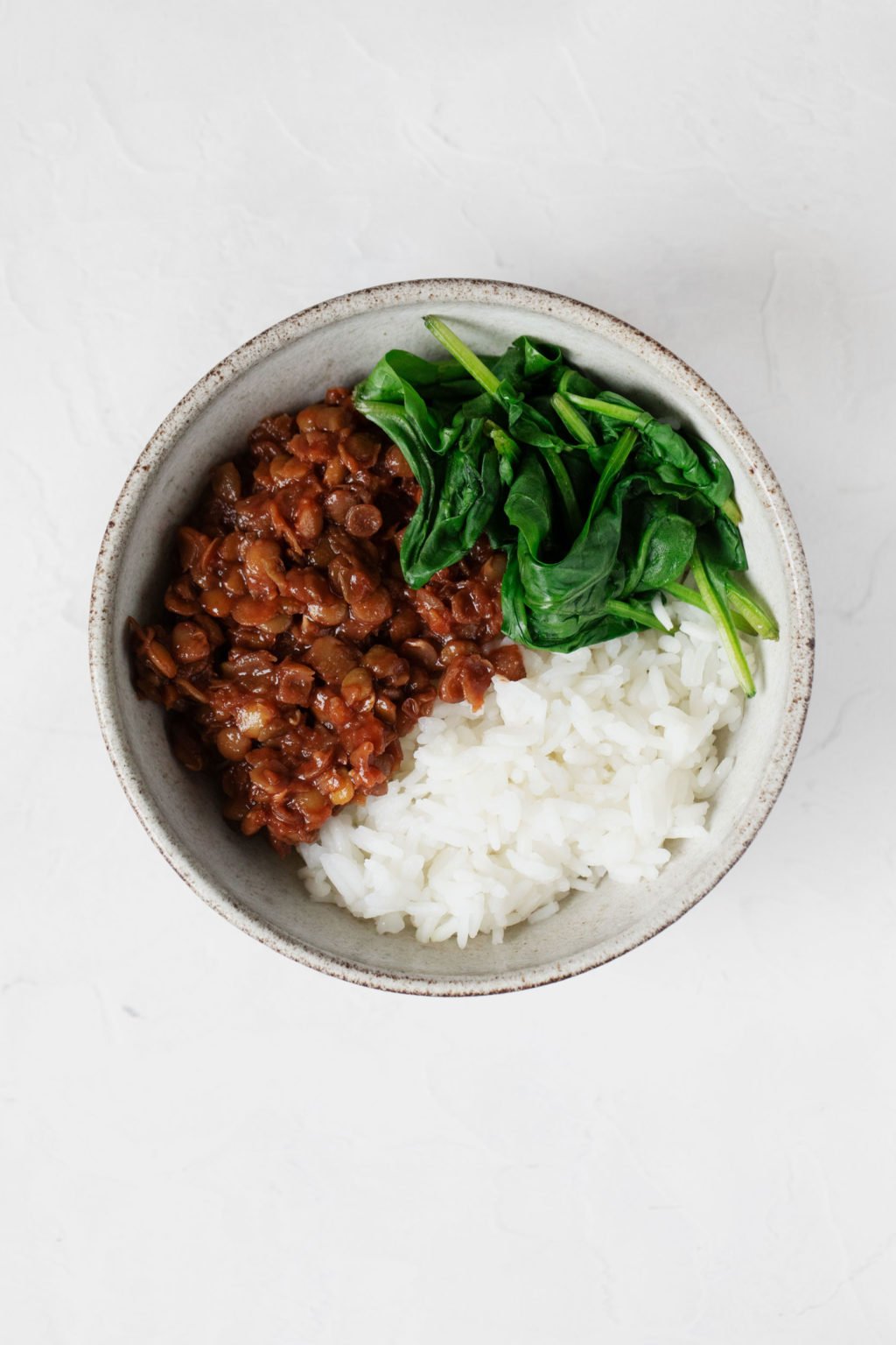 A white and gray hued bowl is filled with legumes, rice, and steamed baby spinach.