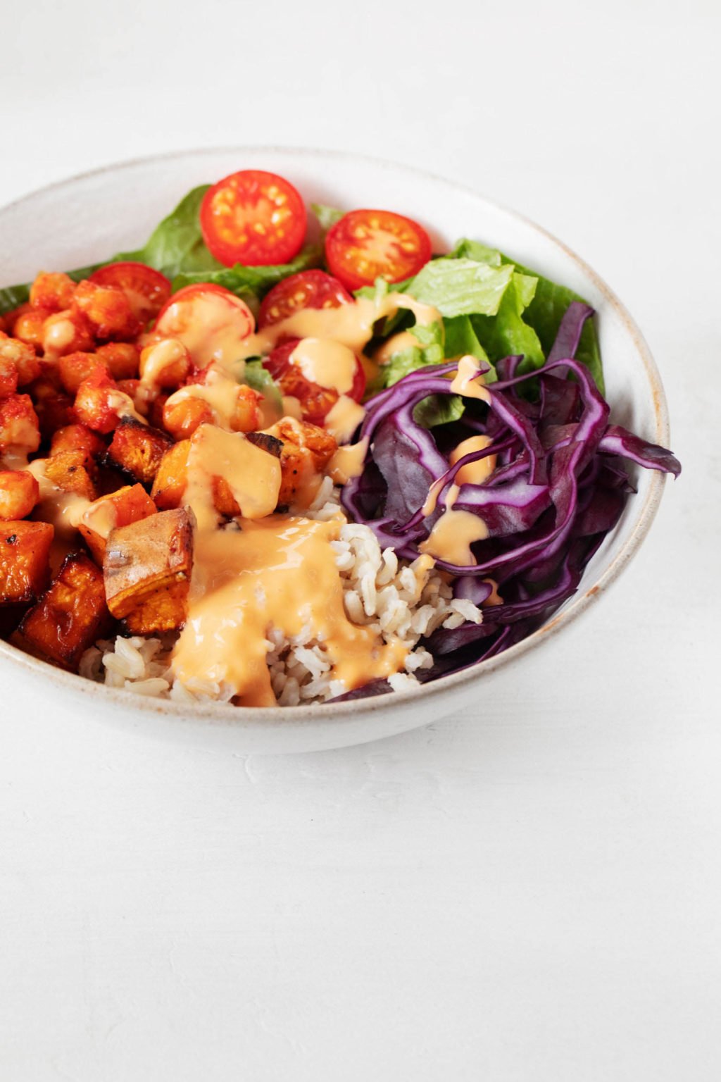 An angled photograph of a white ceramic bowl, resting on a white surface. The bowl has been piled with chickpeas, roasted sweet potatoes, colorful vegetables, and a creamy sauce.