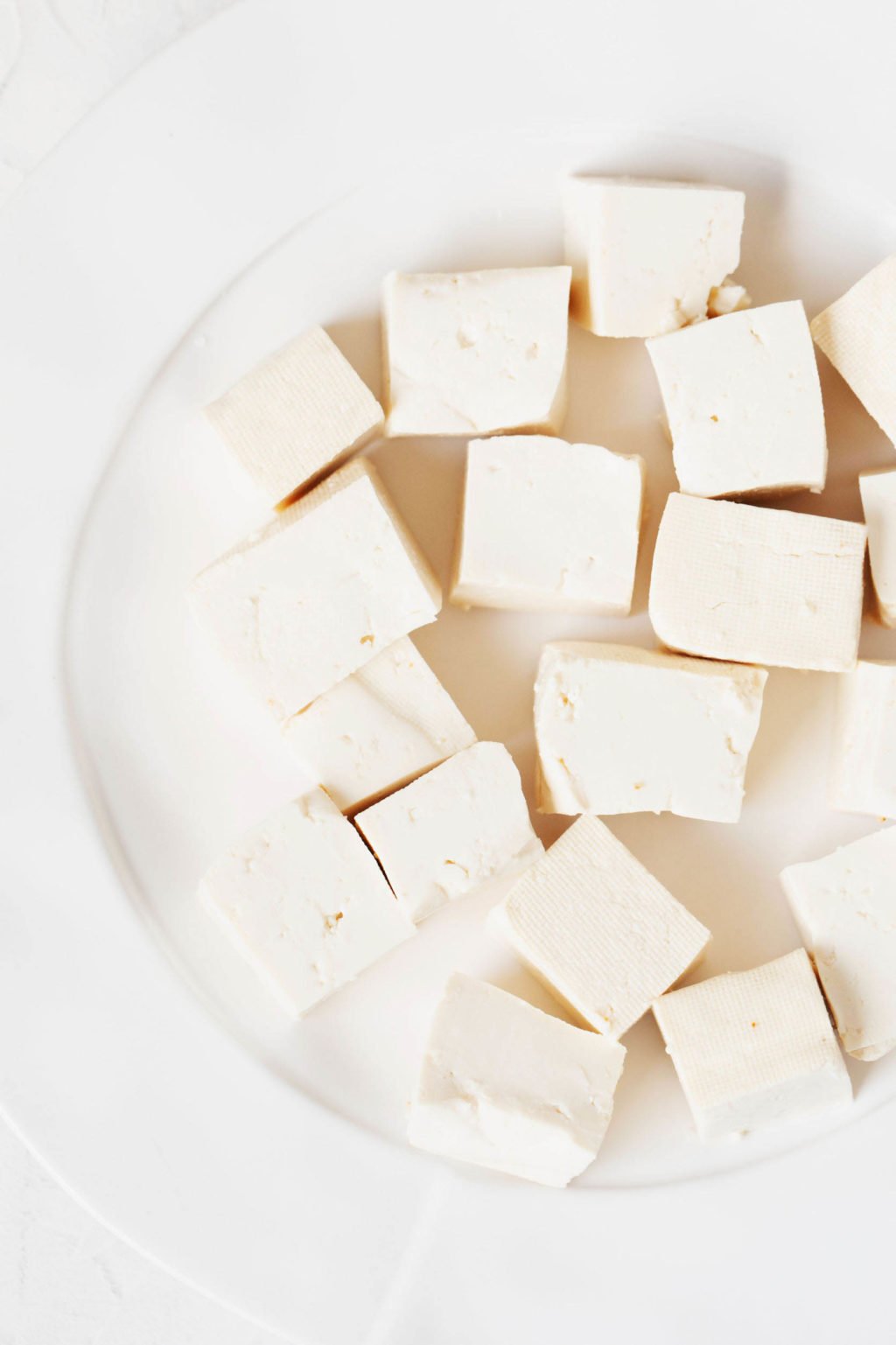 Cubed, plain tofu is resting on a white plate. The plate is on a white surface.
