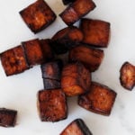 Deep, reddish brown cubes of baked balsamic tofu are resting on a white surface.