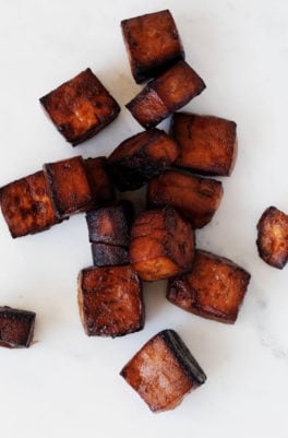 Deep, reddish brown cubes of baked balsamic tofu are resting on a white surface.