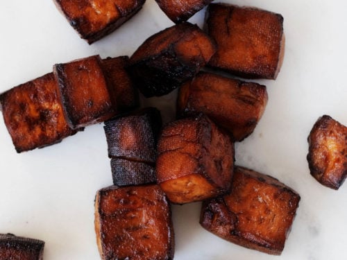 Deep, reddish brown cubes of baked balsamic tofu are resting on a white surface.