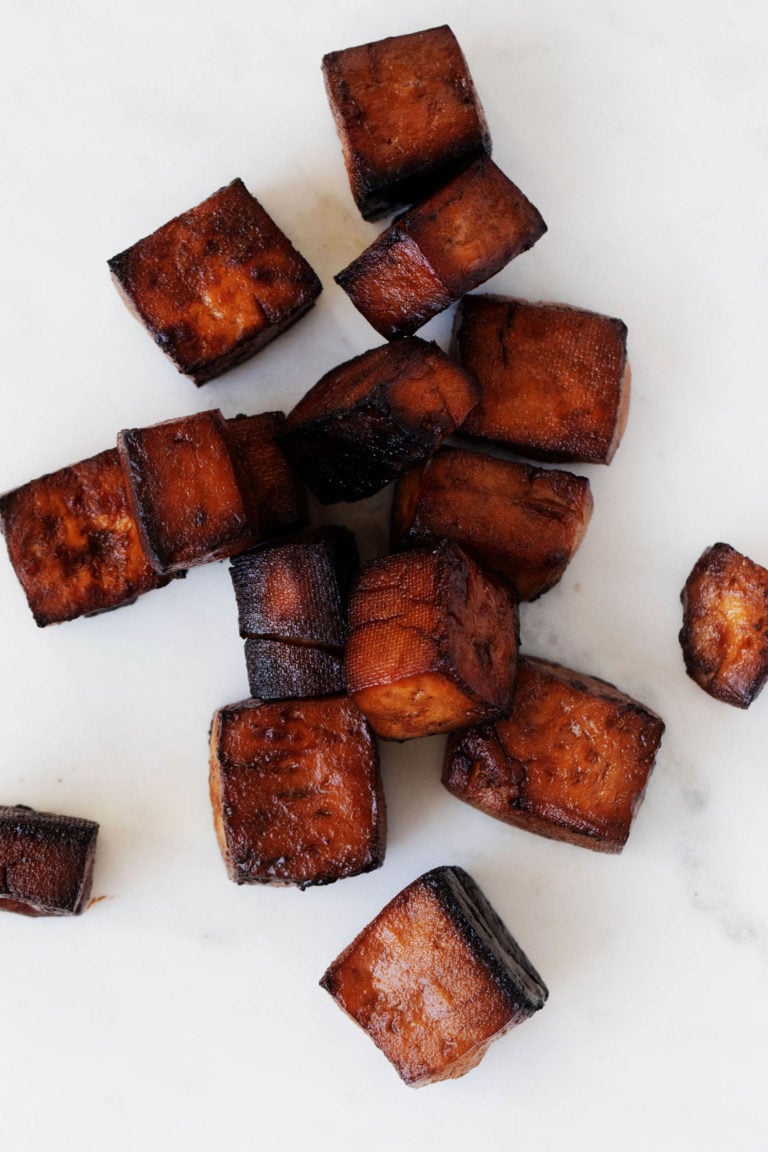 Deep, reddish brown cubes of baked balsamic tofu are resting on a white surface.