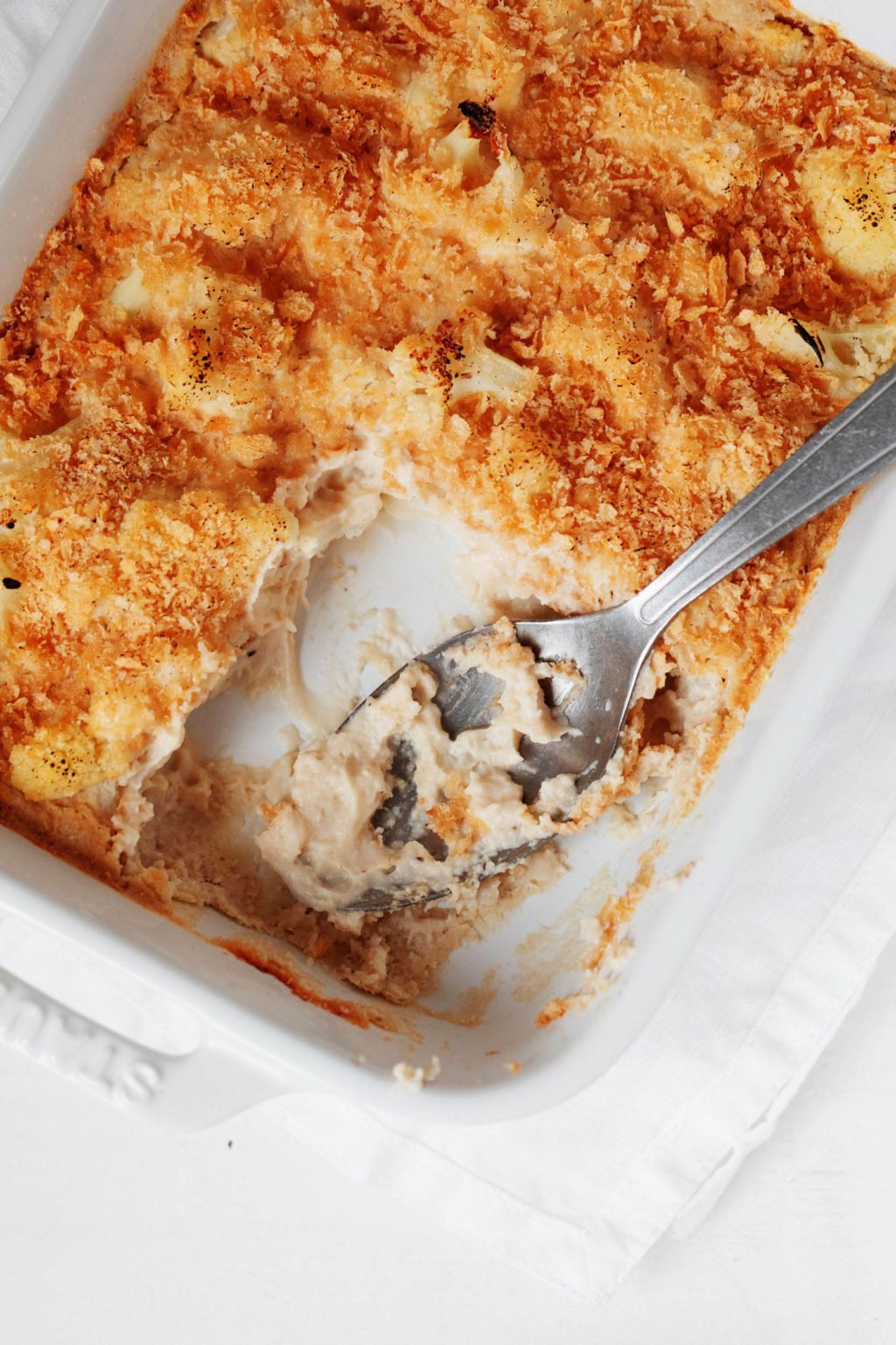 A baked casserole dish is held in a rectangular white pan. It's covered in bread crumbs and being served with a large, silver spoon.