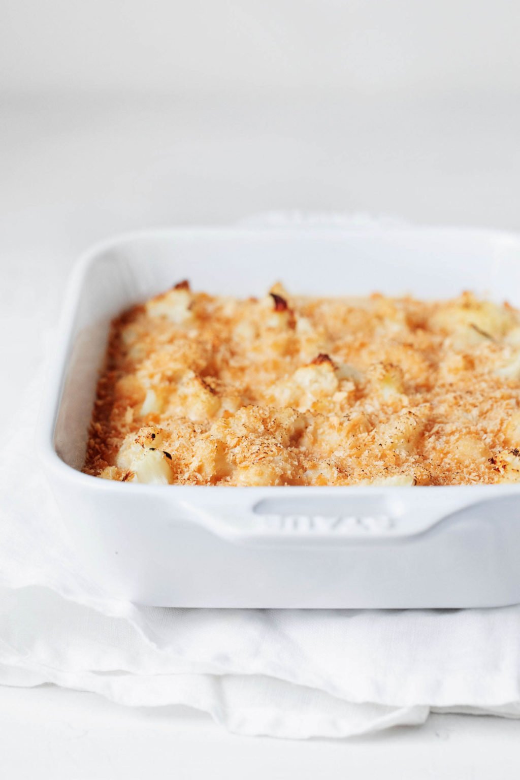 A white, rectangular baking dish has been filled with a cauliflower gratin and topped with breadcrumbs. It rests on a white cloth against a white surface.