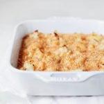 A white, rectangular baking dish has been filled with a cauliflower gratin and topped with breadcrumbs. It rests on a white cloth against a white surface.