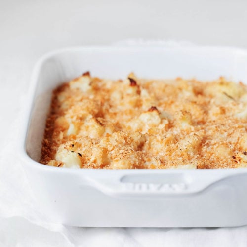 A white, rectangular baking dish has been filled with a cauliflower gratin and topped with breadcrumbs. It rests on a white cloth against a white surface.