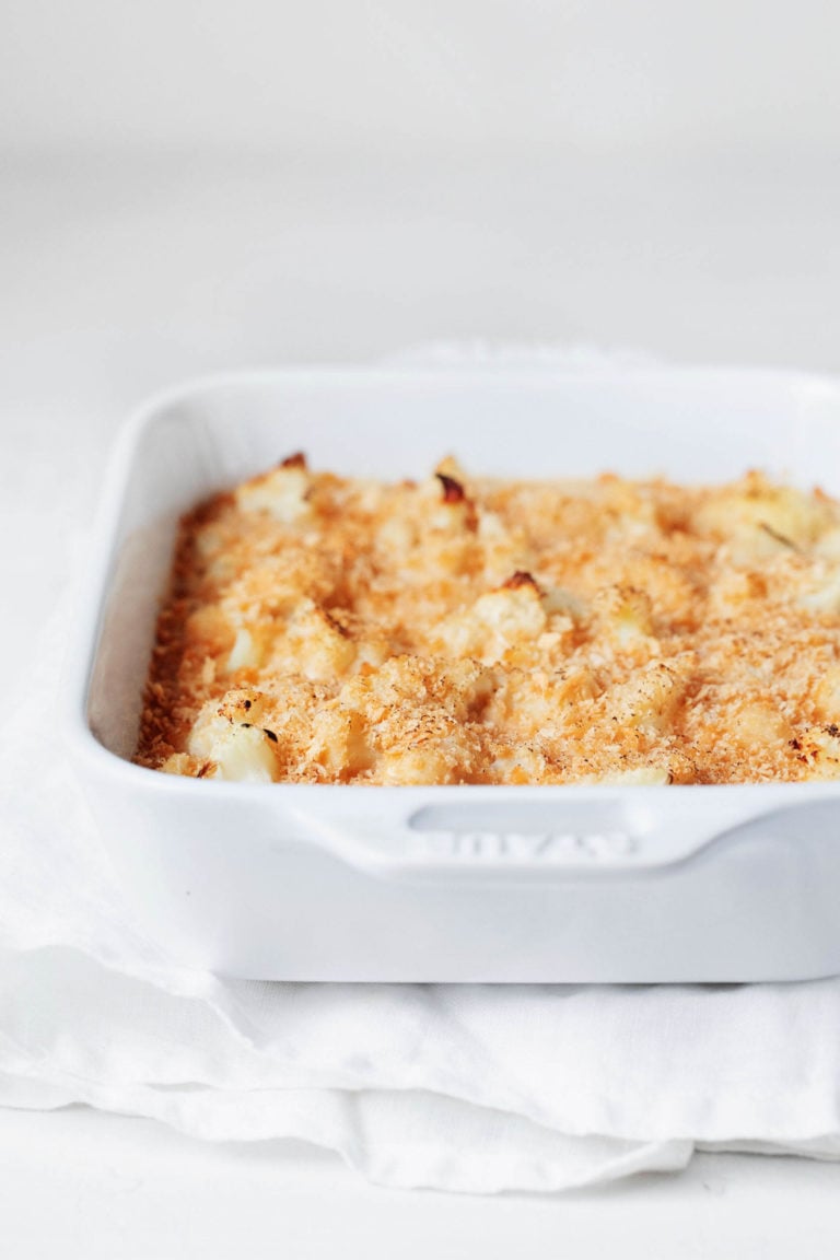 A white, rectangular baking dish has been filled with a cauliflower gratin and topped with breadcrumbs. It rests on a white cloth against a white surface.