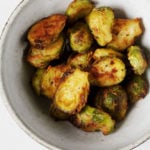 A white, ceramic bowl is filled with glazed miso butter Brussels sprouts. It rests on a white surface.