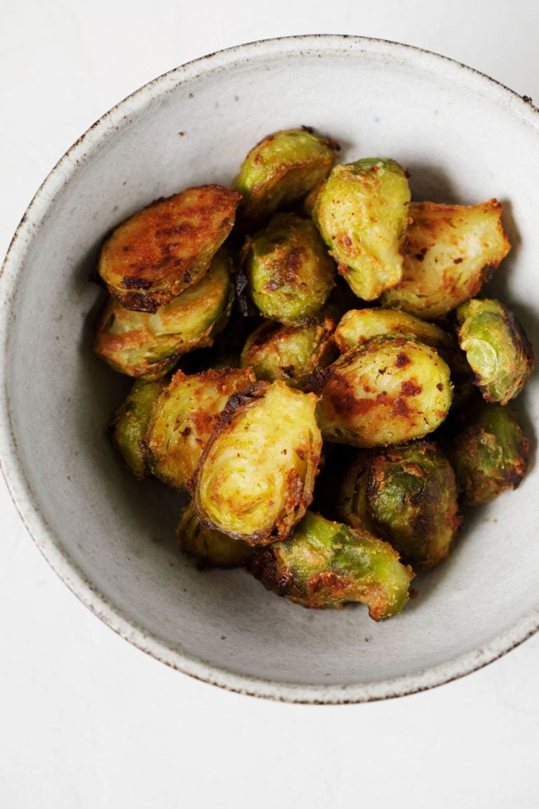 A white, ceramic bowl is filled with glazed miso butter Brussels sprouts. It rests on a white surface.