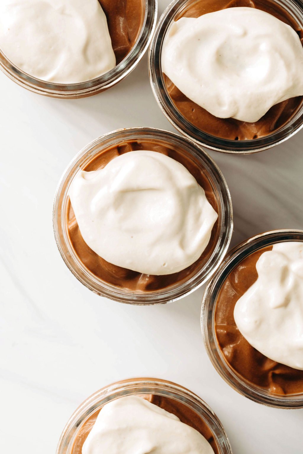 An overhead image of five small glass mason jars, each of which is filled with chocolate pudding and a vegan tofu whipped cream.