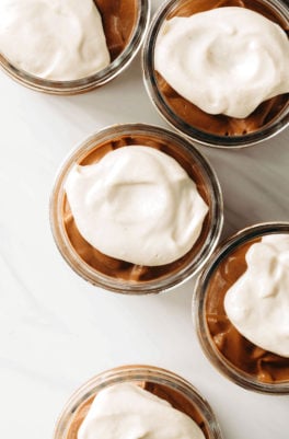 An overhead image of five small glass mason jars, each of which is filled with chocolate pudding and a vegan tofu whipped cream.