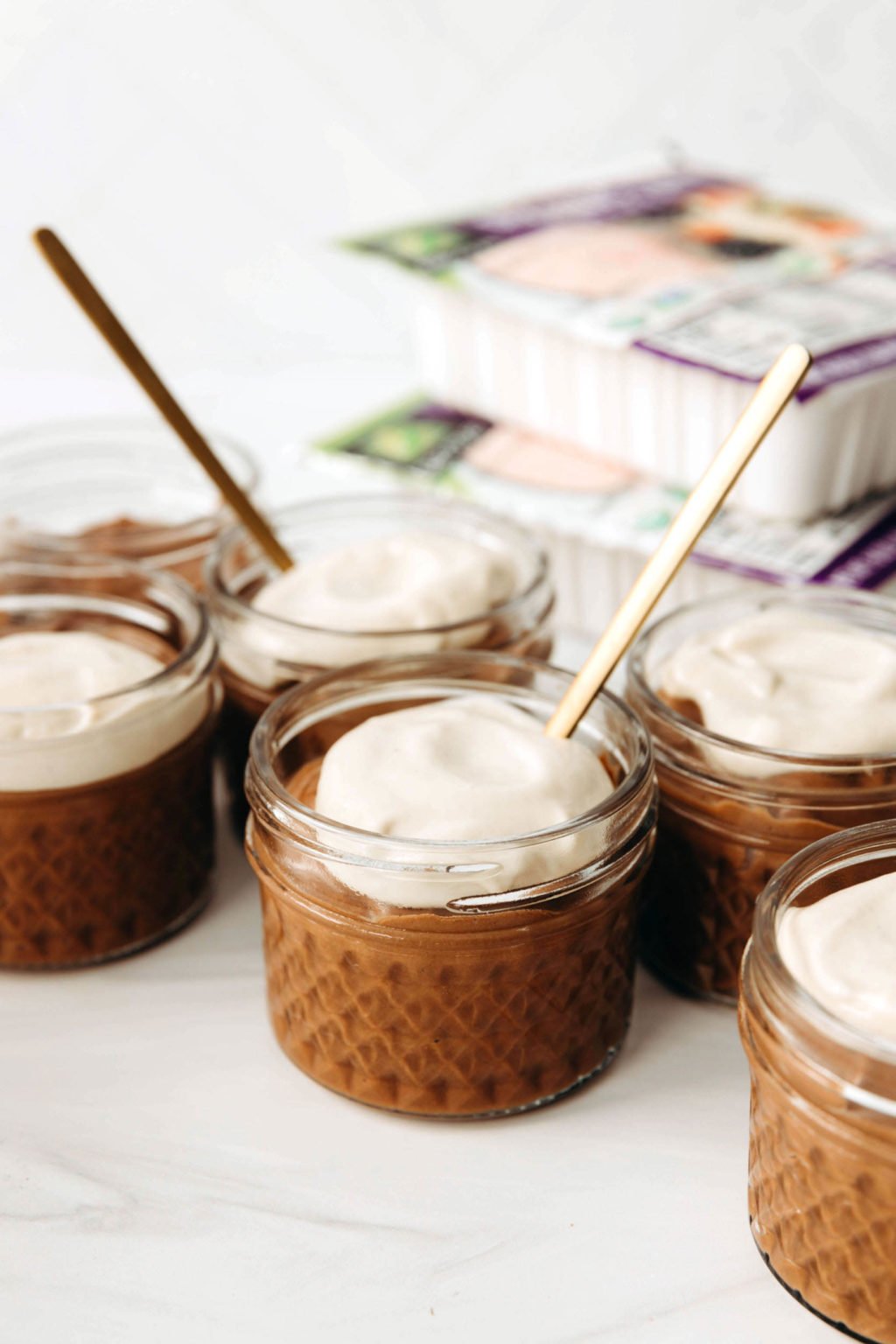 A number of small, round glass jars have been filled with chocolate pudding and a dollop of white cream. Two golden spoons are sticking out from two of the jars.