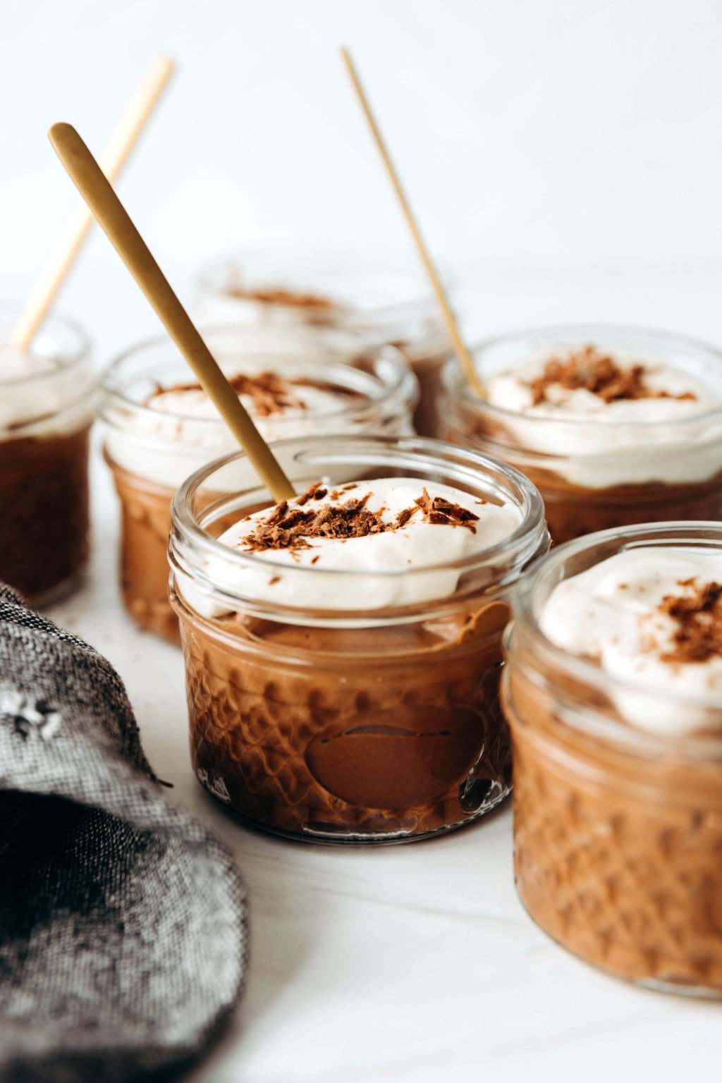 Several small mason jars are resting on a white surface. Each is filled with chocolate pudding and a fluffy tofu whipped cream. They're being served with golden spoons.