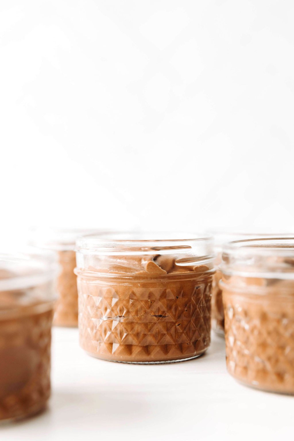An angled photograph of several small, clear glass jars, each filled with chocolate pudding.