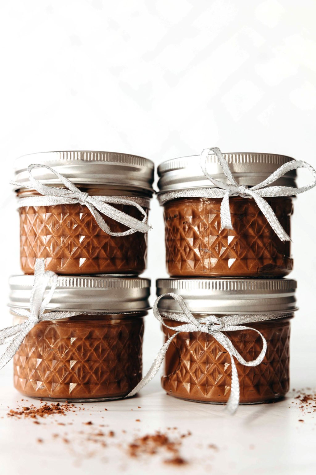 Small, round mason jars have been filled with a chocolate pudding. The jars are stacked against a white backdrop. Each is tied with a festive silver ribbon.