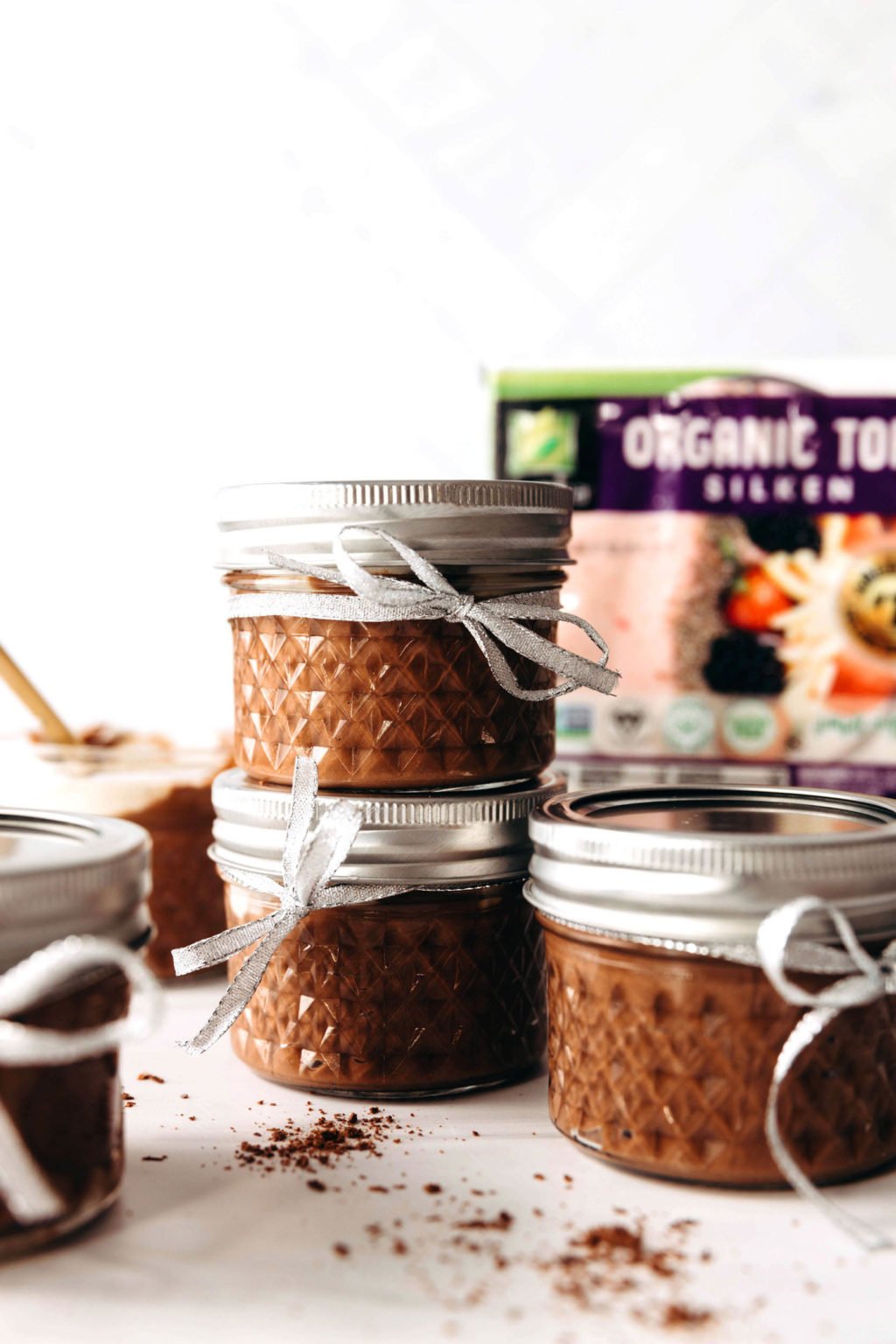 Small mason jars are stacked upon each other on a white surface. They're filled with chocolate pudding and tied with a silver ribbon. Two packages of silken tofu are pictured in the background.