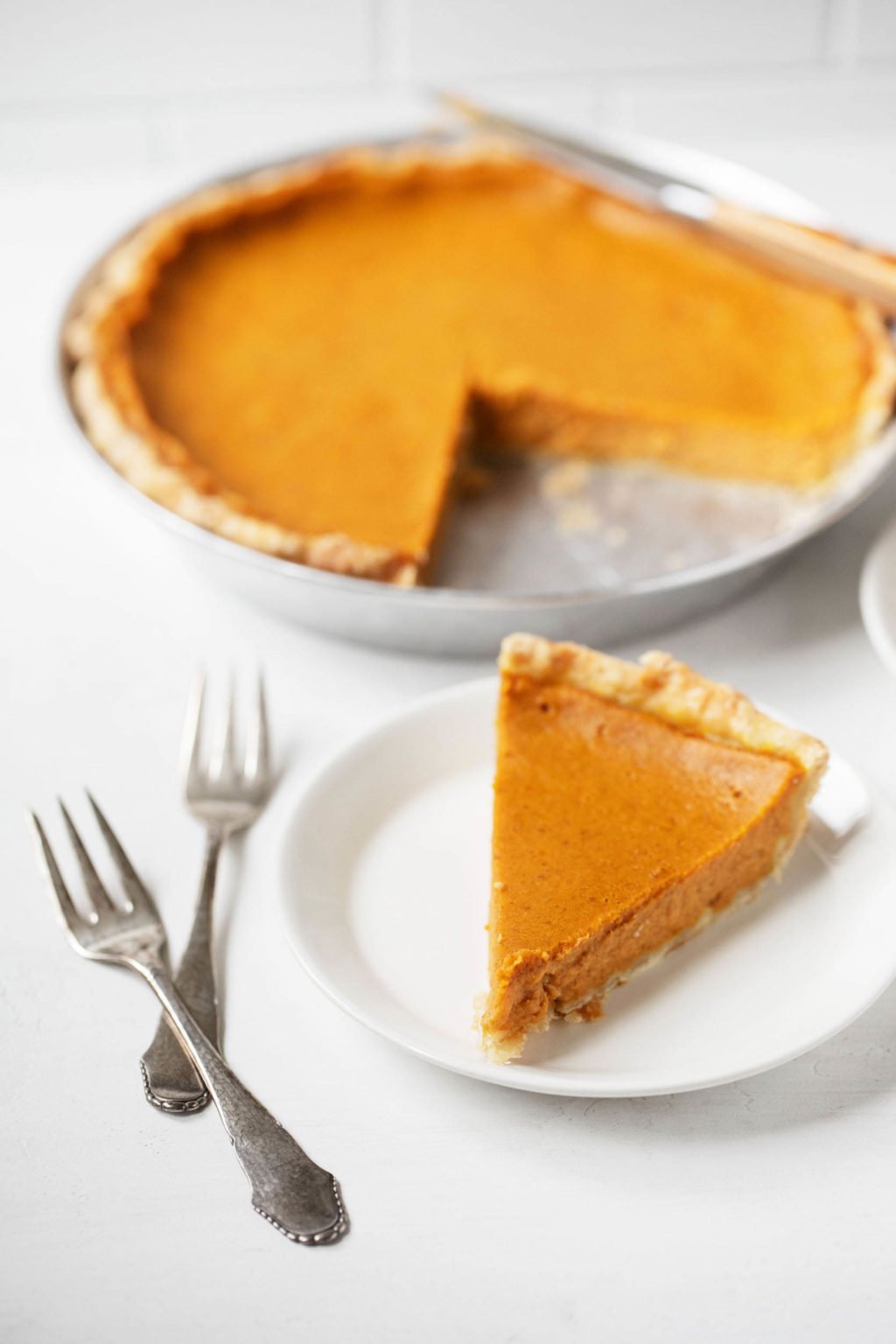 Two silver forks rest on a white surface near a pie plate. A small dessert plate holds one slice of pie in the forefront.